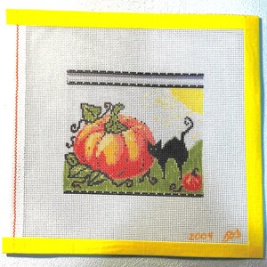 May include: A square needlepoint canvas featuring a cross-stitched design of a large orange pumpkin and a small black cat on a green patch. The background includes a yellow sun and a white sky. The canvas edges are bound with bright yellow tape.