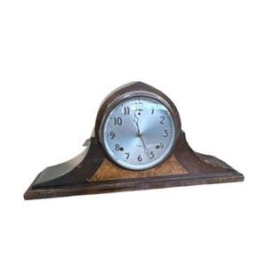 May include: A mantel clock with a dark brown wooden base and a rounded top section. The clock face is silver-toned with black Arabic numerals from one to twelve, featuring two thin metal hands and a glass cover set within a circular metal frame.