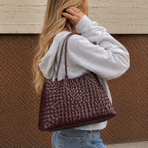 May include: A deep burgundy tote bag featuring an intricate woven pattern across the entire exterior surface. The bag includes two thin, braided shoulder straps attached to the top edge, providing a structured yet flexible rectangular shape.