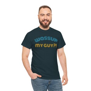 May include: A dark navy blue short-sleeved crew neck t-shirt featuring the text "WASSUP" in light blue and "MY GUY?!" in yellow. The lettering is stylised with a multi-line striped effect, centred on the chest of the garment.