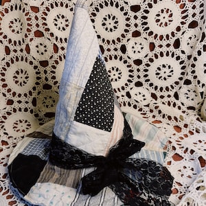 May include: A conical hat constructed from various quilted fabric patches in shades of blue, white, black, and grey. The hat features a wide brim and a pointed crown, accented with a black lace ribbon tied in a bow around the base of the crown.