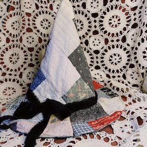 May include: A conical hat constructed from a patchwork of various fabric squares in shades of blue, white, black, and red. The hat features a black velvet ribbon tied in a bow around the base, with visible quilted stitching throughout the fabric.