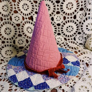 May include: A quilted cone-shaped hat featuring a solid pink fabric crown and a wide brim constructed from a patchwork of blue, white, and floral patterned fabric squares. A dark red frayed ribbon is tied around the base of the crown.