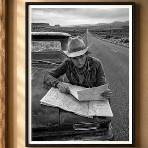 May include: A black and white framed wall art print depicting a person wearing a cowboy hat and a jacket, leaning over the hood of a vintage pickup truck while examining a large paper map on a desolate desert road with distant mountains.