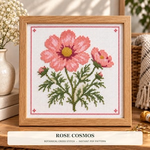 Op de afbeelding: Een ingelijst kruissteekpatroon met een roze cosmosbloem met een geel hart, groene bladeren en een kleine knop. Het ontwerp is geborduurd op witte stof met een roze rand en bevat de tekst ROSE COSMOS BOTANICAL CROSS STITCH.