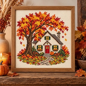 May include: A framed cross-stitch embroidery depicting a white cottage with a red door, green shutters, and a chimney, situated beneath a large tree with vibrant red, orange, and yellow autumn leaves, with a pumpkin near the front entrance.