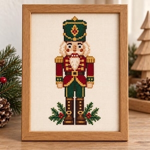 May include: A framed cross-stitch embroidery featuring a nutcracker soldier in a green, red, and gold uniform. The figure wears a tall green hat, a red jacket with gold trim, and dark boots, standing above green holly leaves with red berries.