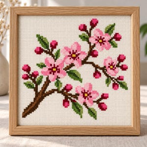 May include: A square cross-stitch embroidery featuring a cherry blossom branch with pink flowers, dark pink buds, and green leaves on white fabric, encased in a light-toned wooden frame. The design displays a brown branch with multiple blooms.