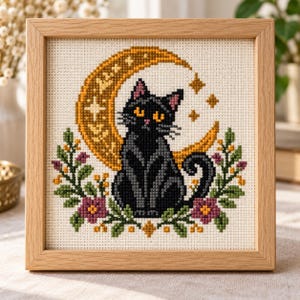 Peut inclure: Une broderie au point de croix carrée dans un cadre en bois clair représentant un chat noir assis devant un grand croissant de lune doré. Le chat est entouré de branches feuillues vertes avec de petites fleurs violettes et des baies jaunes.