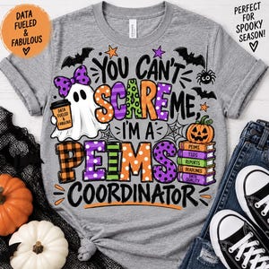 May include: A heather gray t-shirt featuring a graphic with a ghost wearing a purple polka dot bow, holding a coffee cup, and a stack of books topped with a pumpkin. The text reads: You can't scare me, I'm a PEIMS coordinator.
