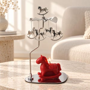 May include: A silver-toned metal candle carousel featuring a rotating top with hanging rocking horse ornaments. A red rocking horse-shaped candle sits on the reflective base, with a lit wick positioned on the back of the horse figure.