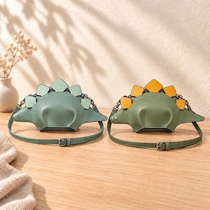 May include: Two dinosaur-shaped crossbody bags with adjustable straps. The left bag is sage green with light green dorsal plates. The right bag is olive green with yellow dorsal plates. Both feature a curved body, pointed sides, and metal hardware.