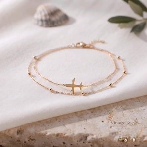 Puede incluir: Una pulsera de tono dorado de doble cadena que presenta un dije de avión en el centro y pequeñas cuentas esféricas a lo largo de los delicados eslabones. La pieza de joyería está diseñada con un cierre y muestra un acabado metálico pulido.