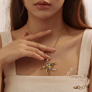 May include: A gold-toned necklace featuring a five-pointed starfish pendant. The starfish has a metallic gold border and a centre inlay of iridescent blue and grey shell material. The pendant hangs from a delicate gold-toned chain.