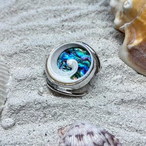 May include: A ring featuring a circular, iridescent abalone shell inlay with a swirling, white, spiral-shaped shell accent set within a polished silver-toned metal band. The center of the inlay displays vibrant shades of blue, green, and purple.
