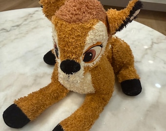 Puede incluir: Un peluche de ciervo con pelaje marrón texturizado, cara de color crema y grandes ojos marrones. El juguete cuenta con pezuñas negras, orejas con puntas negras y una nariz negra, representado en posición sentada con las patas delanteras extendidas.