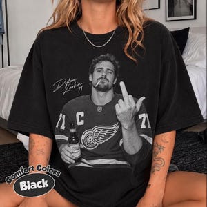 May include: A black short-sleeve t-shirt featuring a black and white graphic of Dylan Larkin wearing a hockey jersey with the number 71, holding a bottle, and gesturing with a middle finger. The text reads Dylan Larkin 71.
