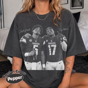 May include: A charcoal grey oversized t-shirt featuring a vintage-style graphic of two football players in jerseys numbered 5 and 17. The text Jayden Daniels and Terry McLaurin is printed above the players, with the word Pepper in the bottom corner.