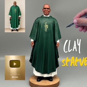 May include: A clay statue of a person wearing a green chasuble with a gold cross emblem over a white alb. The figure has glasses and stands on a circular wooden base. The text CLAY stAtve is visible on the right, and a gold YouTube plaque is on the left.