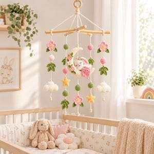 Puede incluir: Un móvil colgante para guardería con una luna creciente de ganchillo color crema con un conejo blanco dormido, rodeado de flores rosas, hojas verdes, estrellas amarillas y nubes blancas de ganchillo suspendidas de un marco de madera.