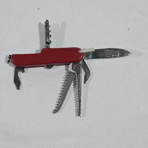 May include: A red multi-tool pocket knife featuring a stainless steel blade, a corkscrew, a bottle opener, a flathead screwdriver, and two serrated saw blades. The handle is solid red with a small white plastic toothpick inserted near the blade.