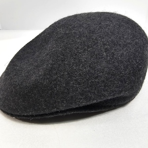 May include: A dark charcoal gray flat cap constructed from a textured, felted wool-like material. The cap features a rounded, structured crown and a short, stiff brim that curves slightly downward at the front, maintaining a classic silhouette.