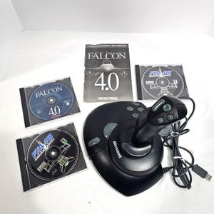 May include: A black Microsoft joystick with a USB cable, accompanied by a Falcon 4.0 communications handbook and three game discs: Falcon 4.0, F/A-18 Hornet, and a second F/A-18 disc, all featuring flight simulation graphics and text.