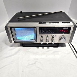 May include: An RCA black and silver portable television and radio combination unit with a small screen displaying static. The device features a digital red clock display, a silver tuning knob, multiple control switches, and a long metal antenna.