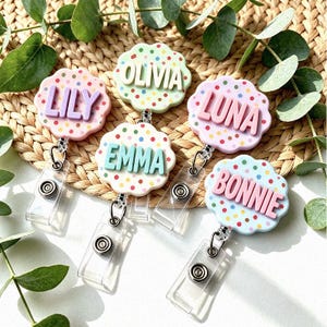 Può includere: Cinque portabadge circolari con bordi smerlati, ognuno caratterizzato da un motivo a coriandoli colorati e lettere in rilievo. I nomi LILY, OLIVIA, EMMA, LUNA e BONNIE sono visualizzati in vari colori pastello sui portabadge.