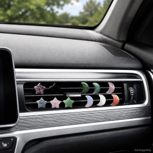 May include: A set of small star and crescent moon shaped decorative clips attached to a horizontal car air vent. The stars are in various colors including purple, black, blue, pink, and green. The moons are green, teal, pink, blue, white, and orange.