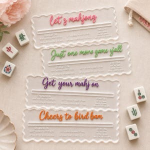 Pode incluir: Quatro suportes retangulares de Mahjong em acrílico transparente com bordas recortadas. Cada suporte apresenta regras impressas de Charleston e Dragões, e uma frase única em escrita colorida: let's mahjong, Just one more game y'all, Get your mahj on e Cheers to bird bam.