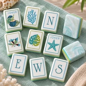 May include: A collection of rectangular Mahjong tiles featuring white faces with blue and green designs, including the letters N, E, W, S, a blue starfish, a green monstera leaf, a blue coral branch, a lotus flower, and a tropical drink.