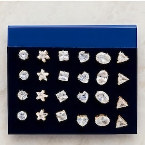 Puede incluir: Una tarjeta de presentación azul oscuro con veinticuatro pendientes de botón dispuestos en cuatro filas de seis. Los pendientes presentan piedras transparentes, facetadas y brillantes en varias formas, incluyendo círculos, estrellas, cuadrados, corazones, óvalos y triángulos.