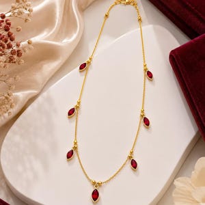 Op de afbeelding: Een gouden ketting met zes gelijkmatig verdeelde, markiesgeslepen rode edelstenen die als bedels hangen. De fijne gouden ketting bevat kleine gouden kraaltjes tussen de zettingen en sluit aan de bovenkant met een karabijnsluiting.
