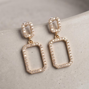 May include: A pair of gold-toned drop earrings featuring two connected rectangular frames. Each frame is encrusted with small, clear, sparkling stones along the entire perimeter. The earrings have a post-back closure with clear plastic earring backs.