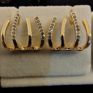 May include: A pair of gold-toned ear cuffs featuring a three-pronged claw design. Each prong is curved, and the outer prongs are embellished with a row of small, clear, sparkling round-cut crystals set along the metal surface.