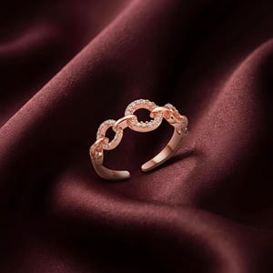 Op de afbeelding: Een open ring in roségoudkleur met een reeks onderling verbonden cirkelvormige schakels. Elke schakel is versierd met kleine, heldere, sprankelende steentjes die langs het buitenoppervlak zijn gezet, wat een verstelbaar kettingontwerp vormt.