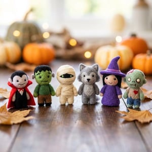 May include: A set of six needle-felted figures including a vampire in a red cape, a green monster, a wrapped mummy, a grey wolf, a witch in a purple hat, and a green zombie with a red heart, all standing in a row on a wooden surface.