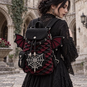 May include: A black and dark red crocheted backpack featuring dragon-like wings on the sides, a central silver spiderweb emblem with a pentagram charm, and a flap closure with a metal snap. The bag has a textured scale pattern and side pockets.
