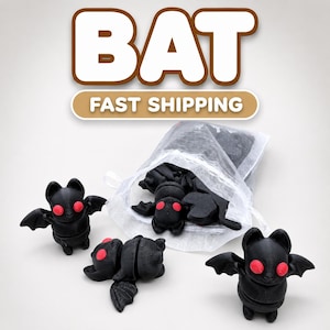 May include: A collection of small black bat figurines with bright red eyes. Two bats stand upright with wings extended, while another lies flat. Several additional bat figurines are contained within a sheer white drawstring organza bag.