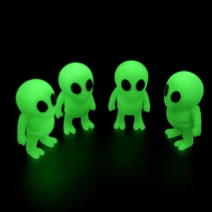 May include: Four small, glow-in-the-dark alien figurines standing in a semi-circle. Each figure features a bright neon green body with a smooth, rounded head, two large oval black eyes, short arms, and two-toed feet, all set against a black background.