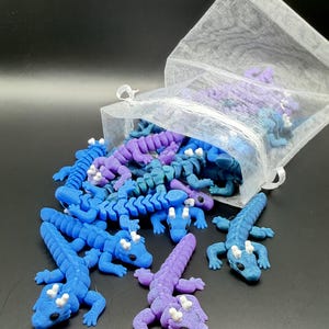 May include: A collection of small, articulated plastic dragon figurines in shades of blue and purple, spilling out of a sheer white organza drawstring bag. Each dragon features a textured body, white horns, and black eyes, with flexible segments.
