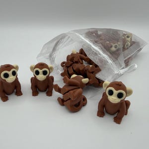 May include: A collection of small brown monkey figurines with cream-colored faces and large black eyes. Three individual monkey figures are positioned in front of a semi-transparent white organza drawstring bag filled with additional monkey figurines.