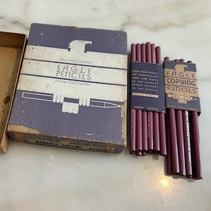 Op de afbeelding: Een vintage paarse kartonnen doos van de Eagle Pencil Company met een adelaarsafbeelding en de tekst Eagle Pencils New York London Toronto. Daarnaast liggen twee bundels kastanjebruine potloden met papieren banden met het opschrift Eagle Copying Pencils.