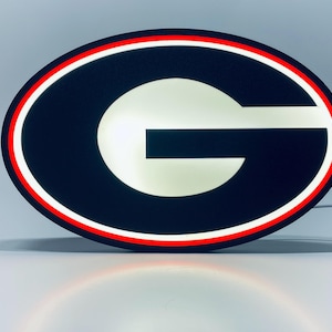 May include: An oval-shaped LED wall sign featuring a large white letter G centered on a black background. The design is enclosed by a thin red border and a bright white illuminated outer ring, creating a high-contrast, glowing display piece.