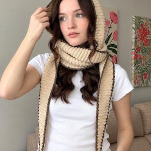 May include: A beige hooded scarf featuring a ribbed knit texture and a dark brown crocheted border. The scarf wraps around the neck and includes a hood that drapes over the head, with two long, pointed ends hanging down the front of a white t-shirt.