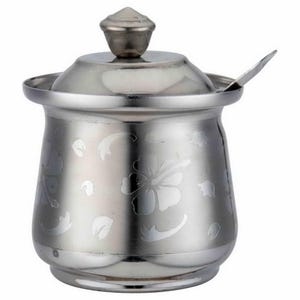 May include: A polished stainless steel sugar bowl with a matching lid and a small spoon. The exterior surface features an etched floral pattern of hibiscus flowers and scattered petals. The container has a rounded body and a knobbed lid.