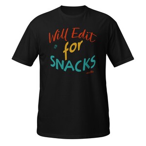 May include: A black short-sleeved t-shirt featuring the text Will Edit for SNACKS in red, yellow, and teal. The design includes a small icon of a coffee cup and a pencil. The lettering is arranged in three lines with a casual, handwritten style.