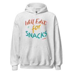 May include: A light gray hooded sweatshirt featuring the text Will Edit for SNACKS in red, yellow, and teal. The design includes a small teal icon of a pill bottle and a small orange icon of a pencil, centered on the front of the garment.
