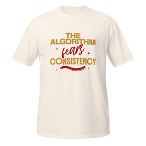 May include: A cream-colored short-sleeve t-shirt featuring the text THE ALGORITHM in gold, the word fears in a red script font, and the word CONSISTENCY in gold. The design includes small red star accents and a red wavy line underneath.